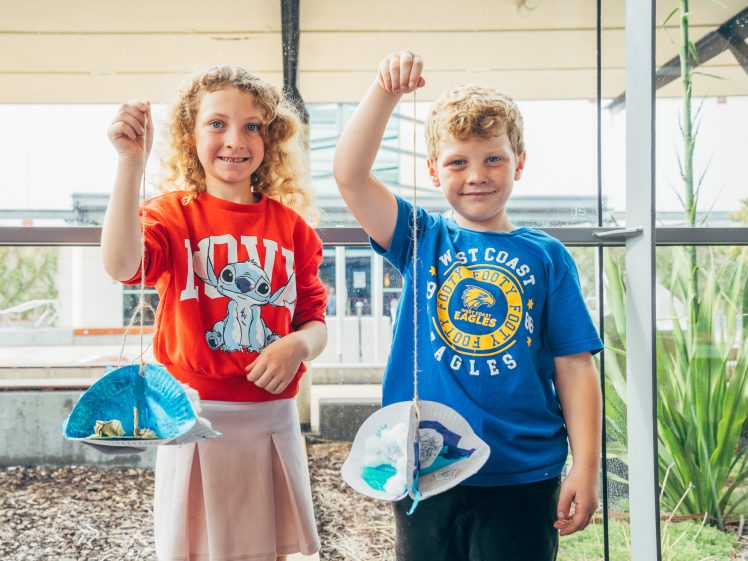 Image of Two smiling children stand indoors holding up arts and crafts projects made from paper plates and string. The girl wears a red shirt with a cartoon animal, and the boy wears a blue West Coast Eagles shirt. Large windows are behind them.