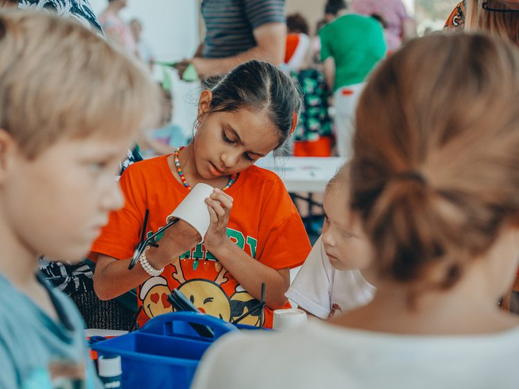 Image of A young girl in an orange shirt decorates a cup with a marker at a crowded craft table, surrounded by other children and art supplies. The scene is lively and colourful.