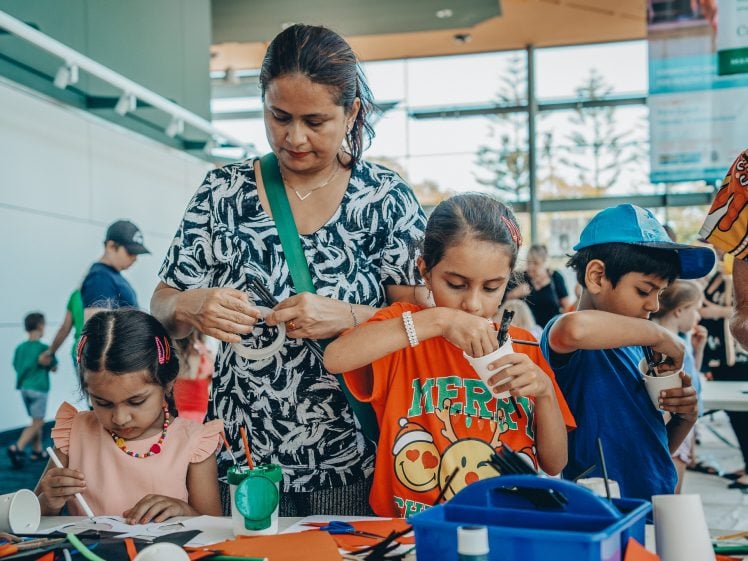Image of A woman stands with three young children at a craft table, helping them with arts and crafts activities. The children are focused on their projects, using paper and other materials inside a bright, indoor community space.