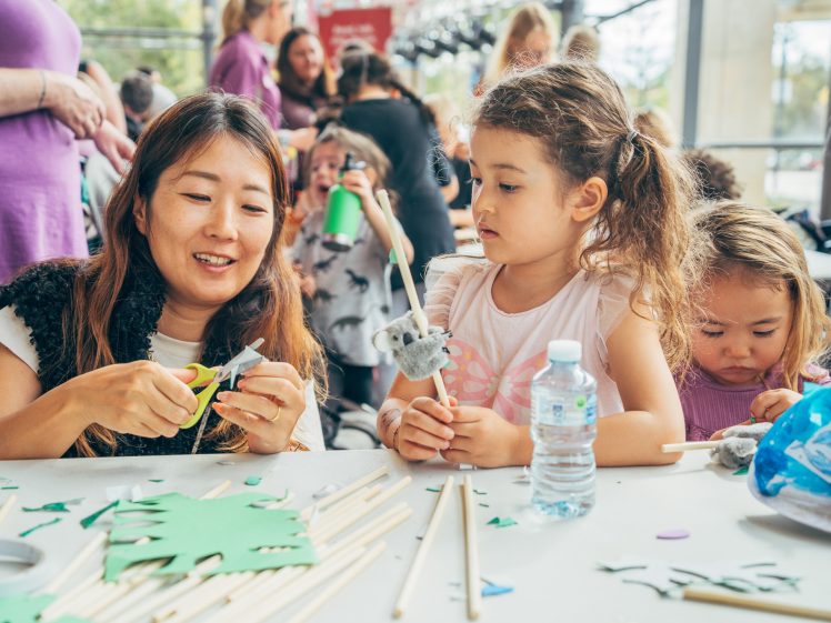 Image of A woman and two young girls sit at a table covered with craft supplies, making animal-themed crafts. The woman is cutting green paper, whilst the girls watch and hold finished crafts. Other people are visible in the background.