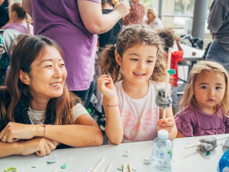 Image of A woman and two young girls sit at a table doing crafts. The girls are smiling, and one holds a handmade craft. The table has supplies and a water bottle. Other people are gathered in the background.