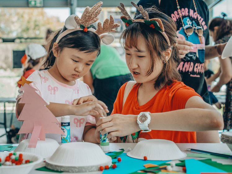 Image of A woman and a child wearing reindeer antler headbands work together on a holiday craft project at a table, surrounded by coloured paper, glue, and decorations.
