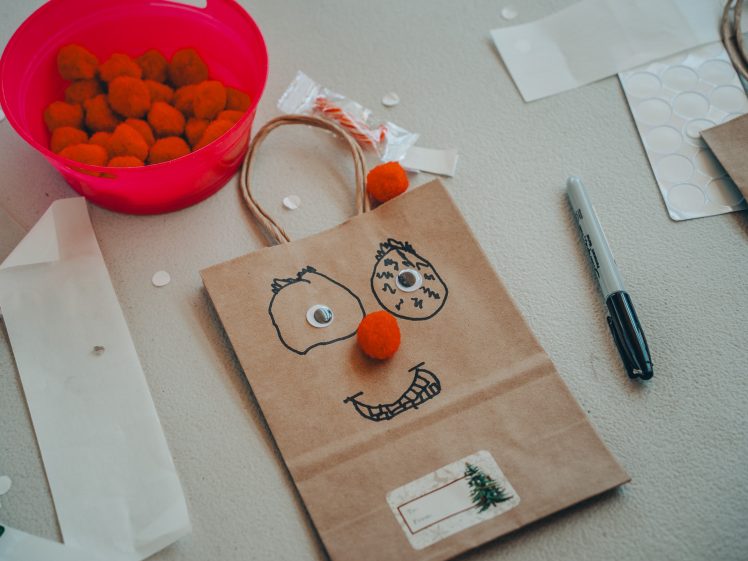 Image of A brown paper bag decorated with a hand-drawn face, googly eye, and orange pompom for a nose sits on a table near a Sharpie marker, stickers, and a pink container of orange pompoms.