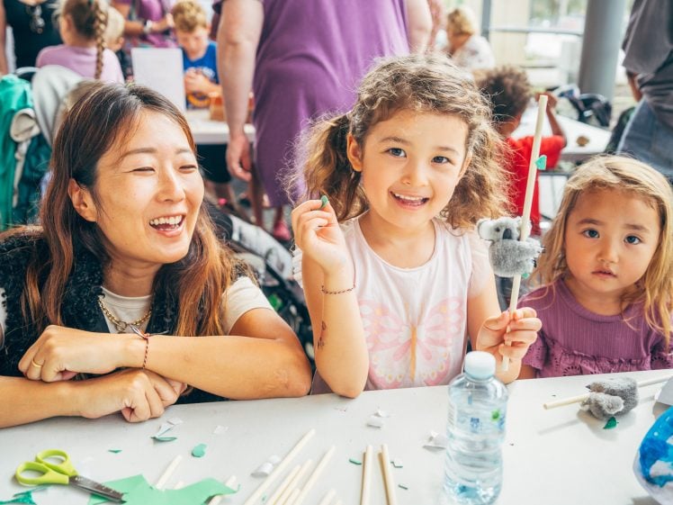 Image of A woman and two young girls sit at a table covered in craft supplies. The girls are smiling, holding handmade koala puppets. The woman is laughing. Other people are visible in the busy, colourful background.