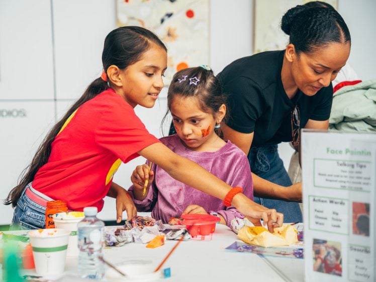 Image of Two young girls and a woman engage in arts and crafts at a table. One girl reaches for supplies while the other, with face paint, watches. The woman, also helping, leans in, and materials are spread out on the table.