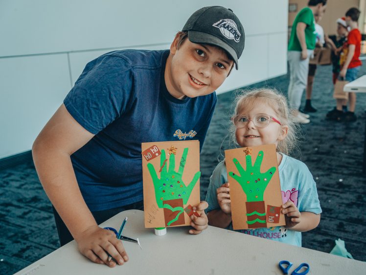 Image of A smiling boy and young girl display handmade crafts featuring green handprints at a table. Art supplies, like scissors and glue, are on the table, and both children look happy and proud of their creations.