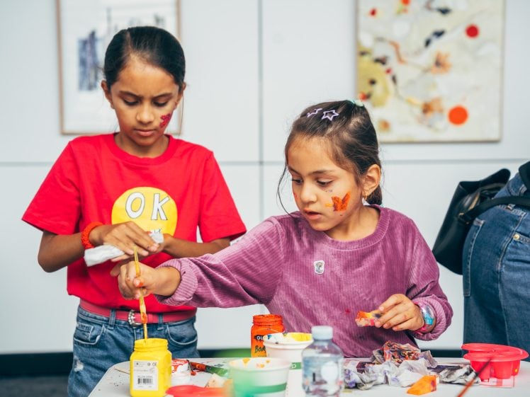 Image of Two young girls paint at a table covered with art supplies. One girl in a red shirt mixes yellow paint, while the other in a purple top paints with orange. Both have face paint and focused expressions. Art is visible in the background.