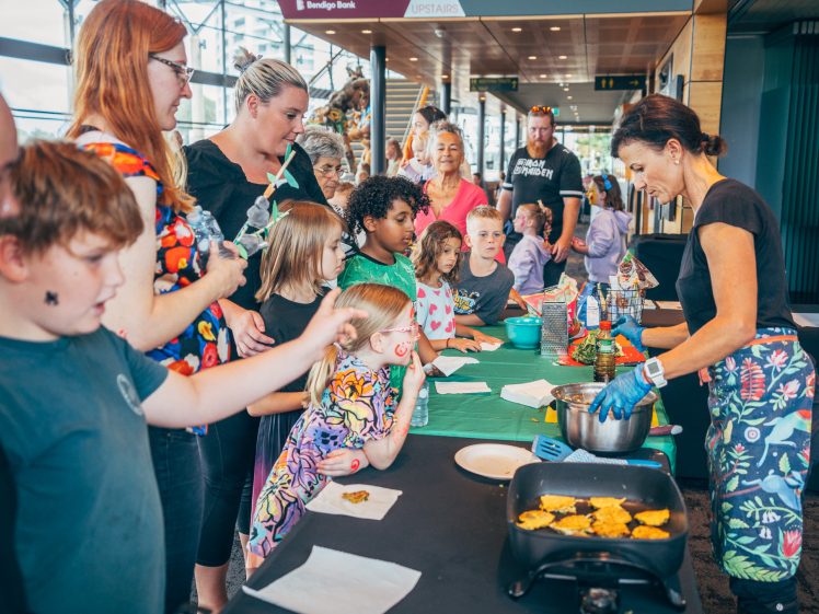 Image of A group of children and adults gather around a table, watching a woman cook food on a hob at an indoor event. The atmosphere is lively, with people eagerly observing the cookery demonstration.
