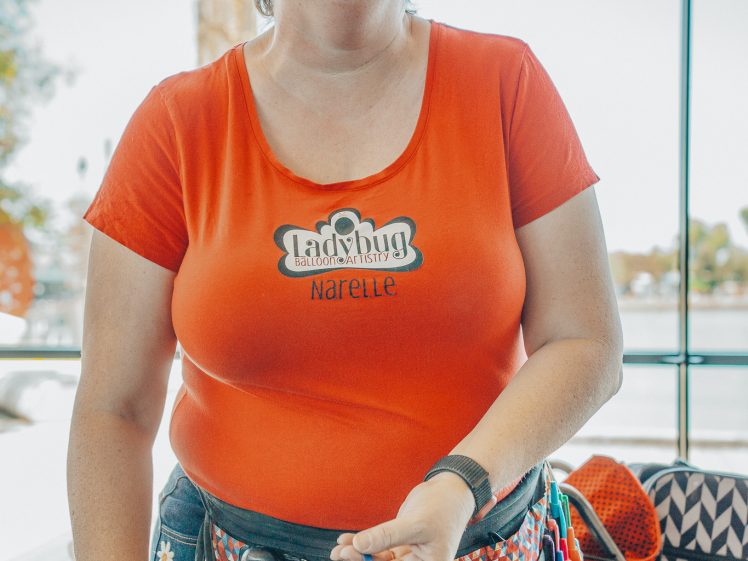 Image of A smiling woman wearing glasses, a red shirt with “Ladybug” and “Narelle” printed on it, and a colourful apron stands indoors. She holds balloon-making tools and has a balloon headband on her head.