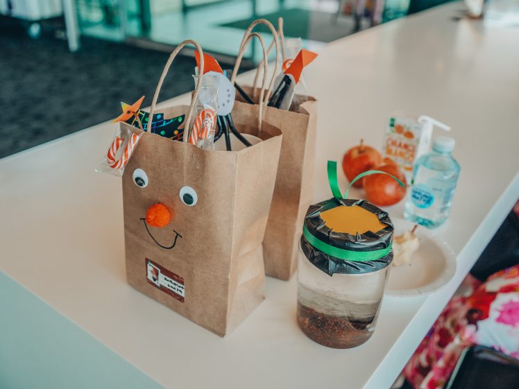 Image of A brown paper bag decorated with googly eyes and an orange pom-pom nose sits on a counter near another gift bag, a jar with a black and green lid, a plate, an apple, hand sanitiser, and a juice carton.