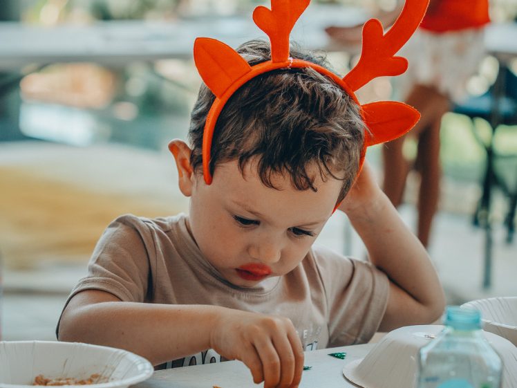 Image of A young boy wearing orange antler headbands concentrates on crafting at a table covered in paper, bowls, and a water bottle. Another child is visible in the blurred background.