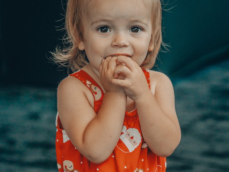 Image of A young child with light brown hair in a top knot wears a red dress with Father Christmas prints, holding their hands near their mouth and looking at the camera with a playful expression.