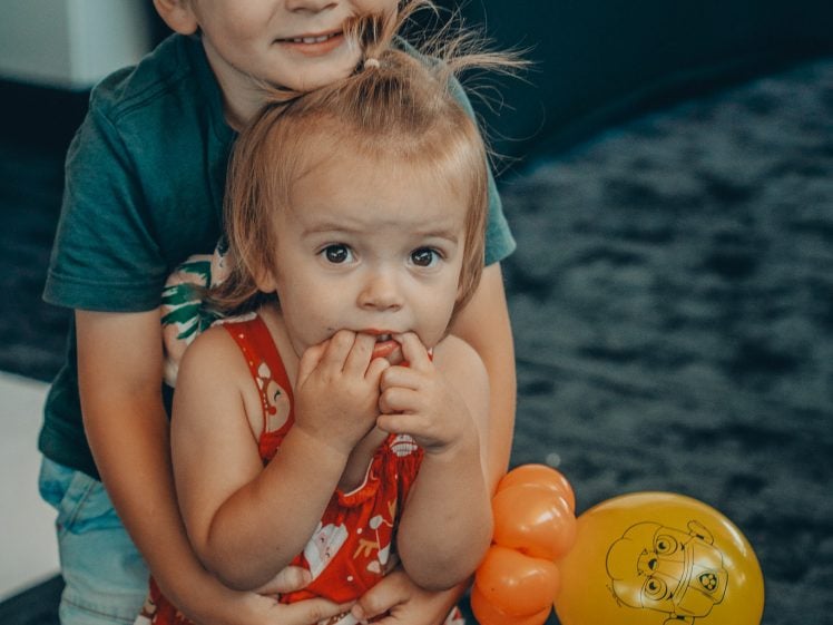 Image of A young boy hugs a younger girl from behind. The girl wears a red outfit with Christmas patterns and holds her fingers near her mouth. A yellow balloon with a cartoon face is tied to her back. Both children stand on a dark carpet.