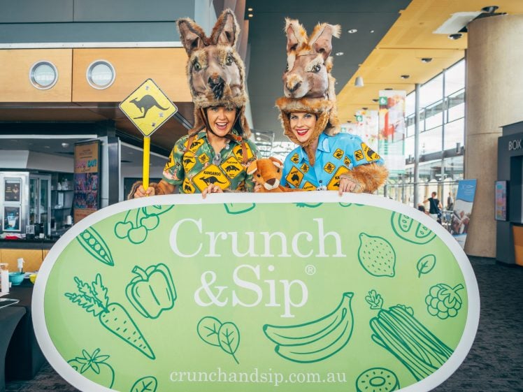 Image of Two people wearing kangaroo costumes and holding a kangaroo crossing sign stand behind a large green Crunch & Sip sign decorated with fruit and vegetable illustrations at an indoor event.