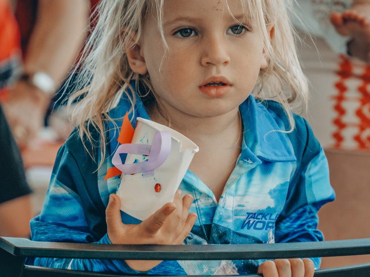 Image of A young child with long blonde hair, wearing a blue shirt, holds a decorated white cup and looks ahead with a thoughtful expression. Adults and other people are blurred in the background.