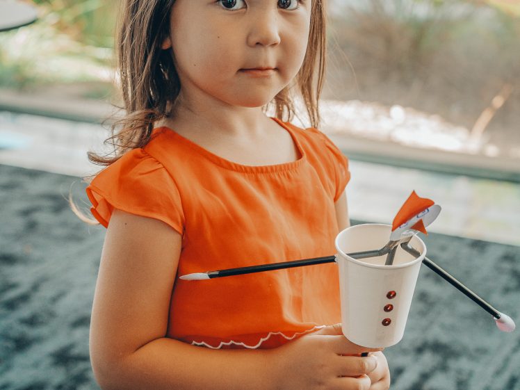 Image of A young girl in an orange dress, wearing a green Christmas tree headband, holds a white cup decorated as a snowman with craft supplies sticking out of it. She stands indoors with a neutral expression.