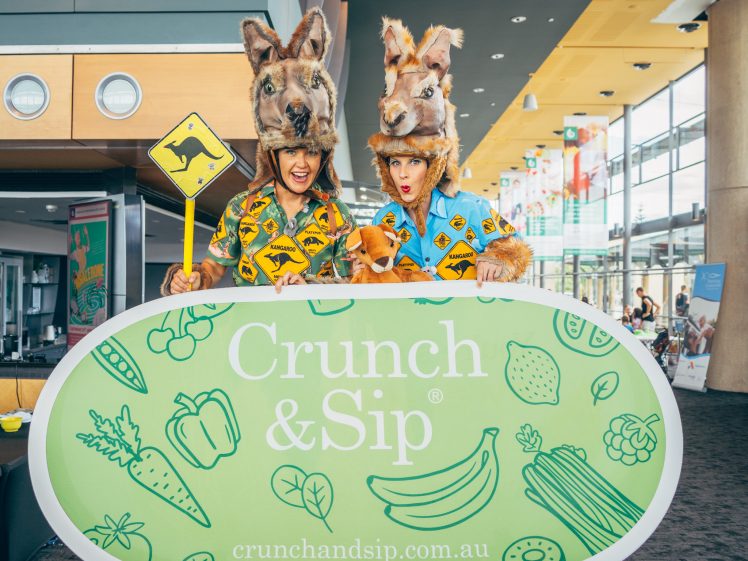 Image of Two people dressed in kangaroo costumes hold a yellow road sign and stand behind a green Crunch & Sip banner decorated with fruit and vegetable illustrations, inside a modern building.