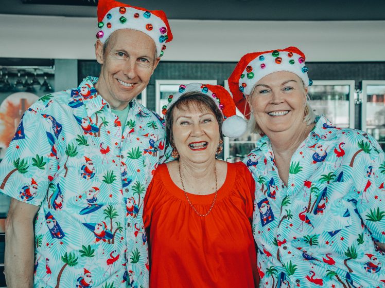Image of Three smiling adults pose together indoors. Two wear festive Father Christmas hats and matching tropical Christmas shirts, while the woman in the middle wears a bright red top. All appear cheerful and celebratory.