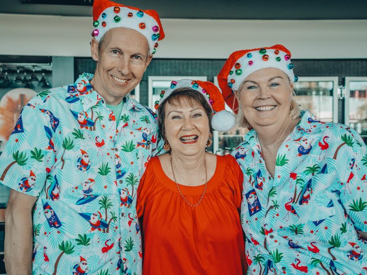 Image of Three adults smile together indoors. Two wear Christmas hats with baubles and matching tropical shirts featuring holiday designs. The woman in the middle wears a red blouse. The scene feels festive and joyful.