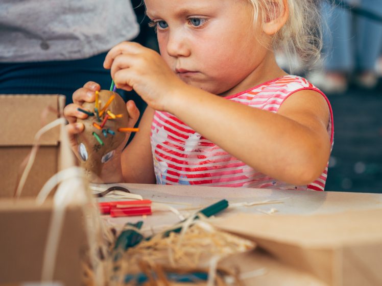 Image of A young girl with blonde hair, wearing a red and white striped shirt, sits at a table and concentrates on assembling a craft project with small colourful parts and straw-like materials in front of her.
