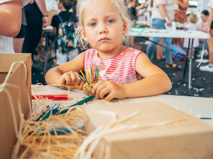 Image of A young girl with blonde hair, wearing a pink striped shirt, sits at a craft table holding coloured pencils, with other people and children working on activities in a busy indoor setting.