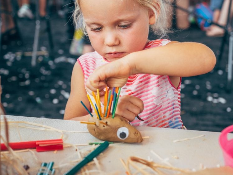 Image of A young girl concentrates as she decorates a potato with colourful sticks and a googly eye at an arts and crafts table with other children and adults in the background.