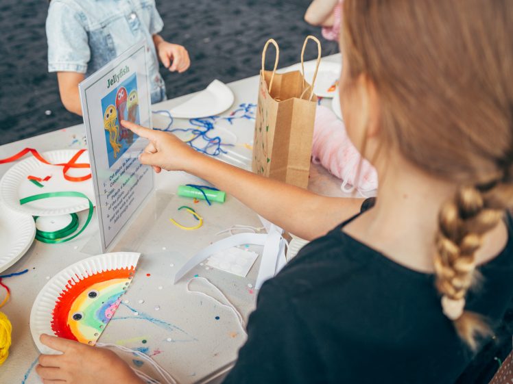 Image of A child with plaited hair points at a jellyfish craft instruction sheet on a table covered with colourful craft supplies, paper plates, and wool, while other children work nearby.