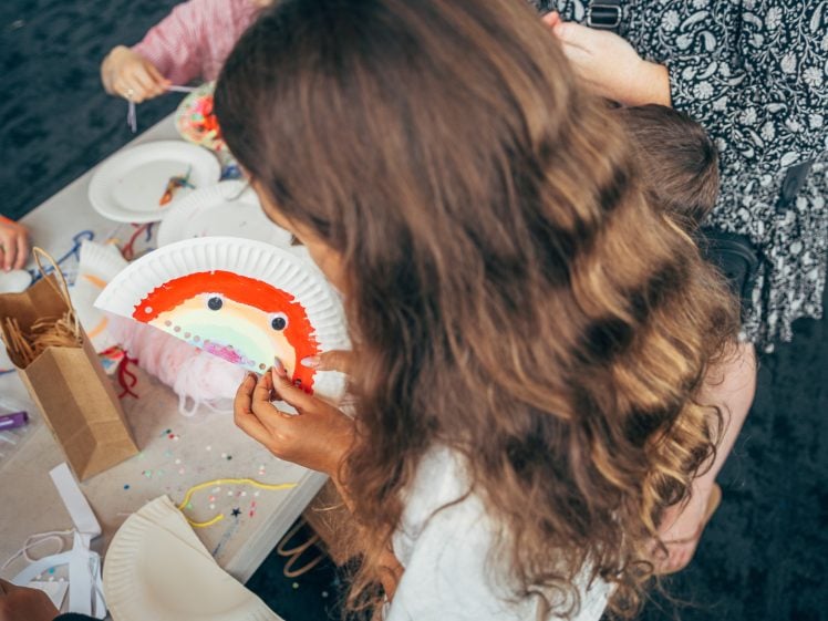 Image of A child with long, wavy hair holds a paper plate painted with a colourful rainbow and googly eyes, while sitting at a table with craft supplies and brown paper bags. Other children and an adult are nearby.