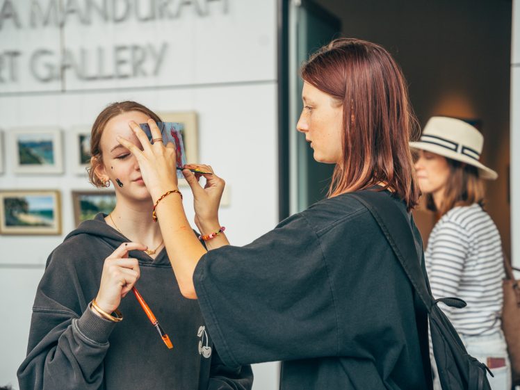 Image of A woman applies face paint to another woman standing outside an art gallery, whilst a third person in a hat and striped shirt walks by in the background. Framed artwork is visible on the gallery wall.