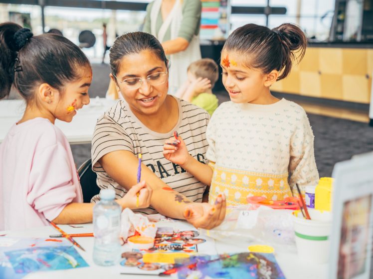 Image of A woman sits at a table with two smiling girls, showing them her hand as they paint and draw together. The girls have face paint and art supplies, and appear engaged and happy in a bright, indoor setting.