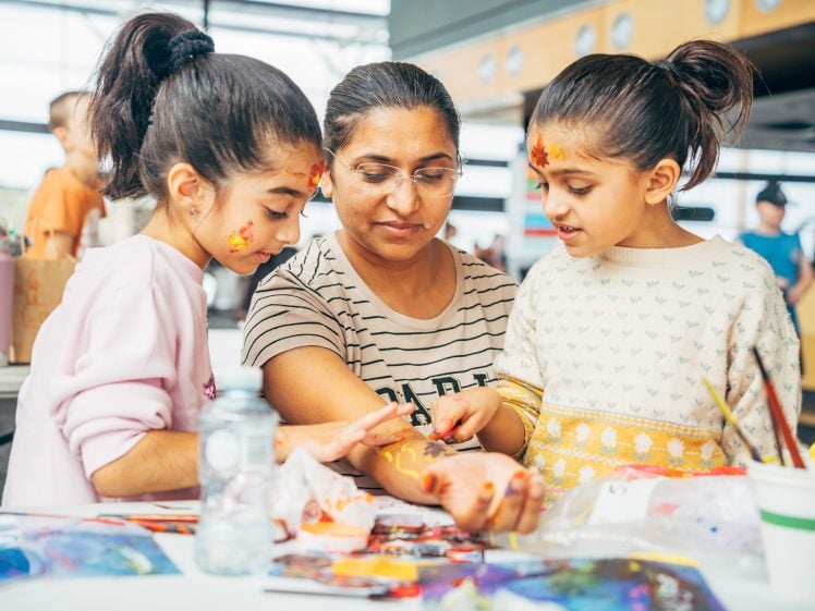 Image of A woman helps two young girls paint their hands at a table filled with art supplies. All three have face paint and are focused on their creative activity in a bright indoor space.