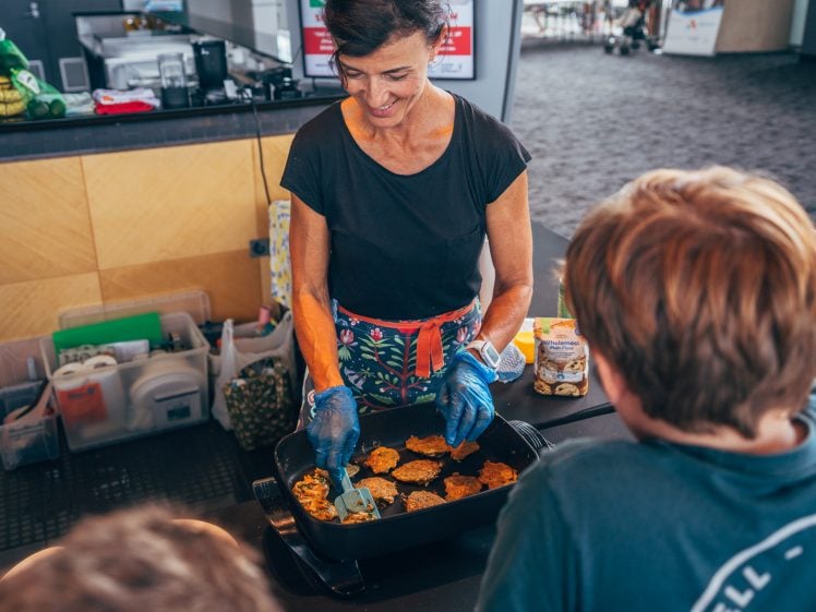 Image of A woman wearing gloves and an apron cooks food on a grill while two children watch. She is smiling and using a spatula, with cooking supplies and ingredients visible on the worktop nearby.