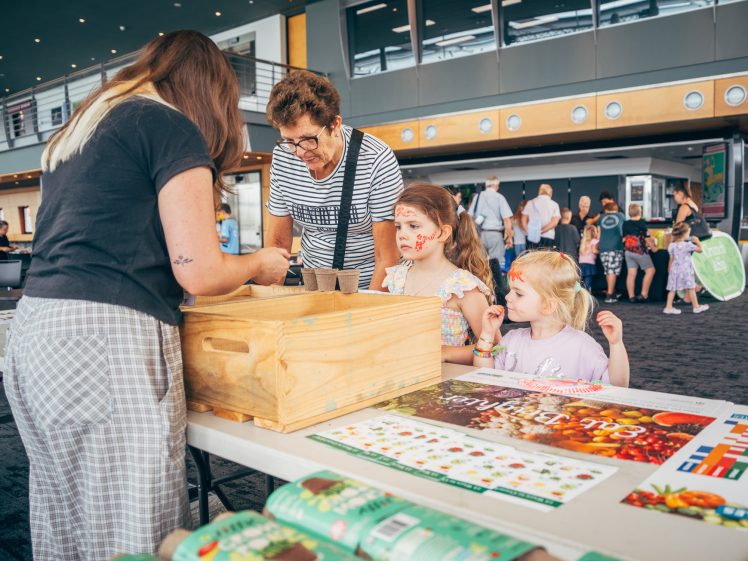 Image of A woman helps two young girls with face paint at a table, whilst another adult watches. The table has a wooden box, posters, and craft materials. Other people are visible in the background of the indoor event space.