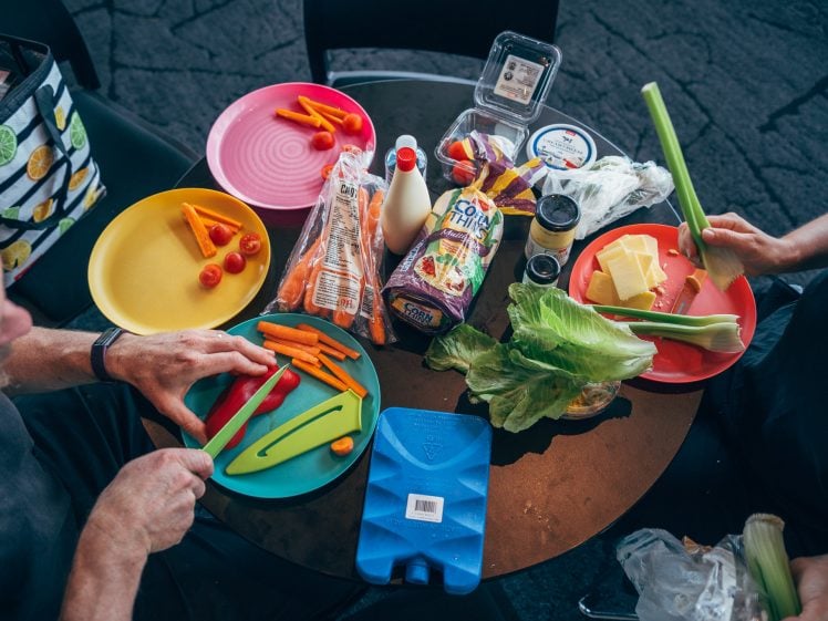 Image of Two people are preparing a meal at a round table with colourful plates, fresh vegetables, bread, cheese, dressing, and snacks. Hands are chopping carrots, celery, and arranging food for a meal.