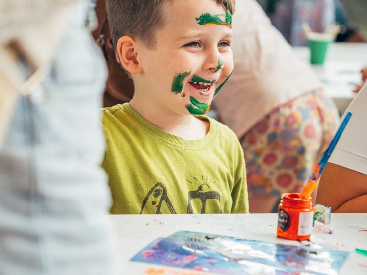 Image of A young boy with green paint on his face smiles whilst sitting at a table covered with art supplies, surrounded by people in a lively, creative setting.