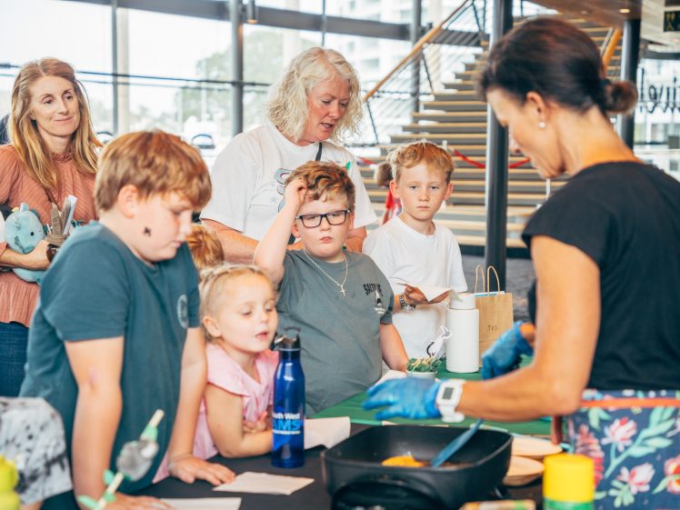 Image of A group of children and two adults watch a woman in gloves demonstrate an activity at a table indoors. The children look curious and engaged, whilst the adults stand nearby observing. There are various items on the table.