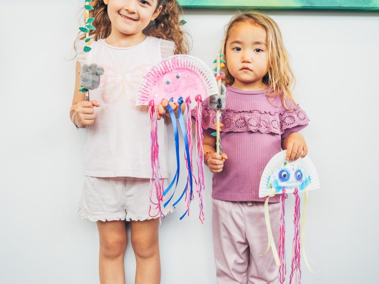 Image of Two young girls stand side by side holding jellyfish crafts made from paper plates, coloured strings, and googly eyes. Colourful paintings of birds are visible on the wall behind them.