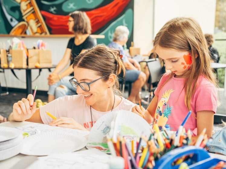 Image of A woman and a young girl do arts and crafts at a table with paper plates, coloured pencils, and drawings. The girl has face paint and both are focused on their artwork. Other people are in the background.