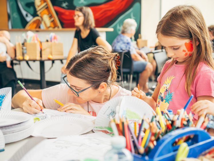 Image of A woman and a young girl with face paint work on art projects at a table covered with coloured pencils, paper plates, and drawings, in a lively room with other people and vibrant wall art in the background.