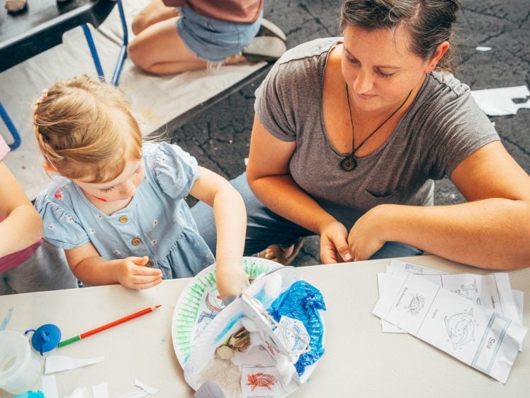 Image of A woman sits beside a young girl who is crafting with paper and art supplies at a table. The girl wears a blue dress and focuses on her project, whilst papers and drawings are spread out around them.