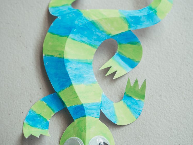 Image of A colourful paper lizard craft painted with blue and green stripes and decorated with two large plastic googly eyes, lying on a light textured surface.