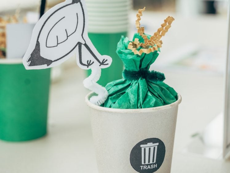 Image of A paper cup labelled RUBBISH contains a green tissue bundle and a paper bird decoration on a pipe cleaner, resembling an ibis picking at rubbish. Green cups are blurred in the background.