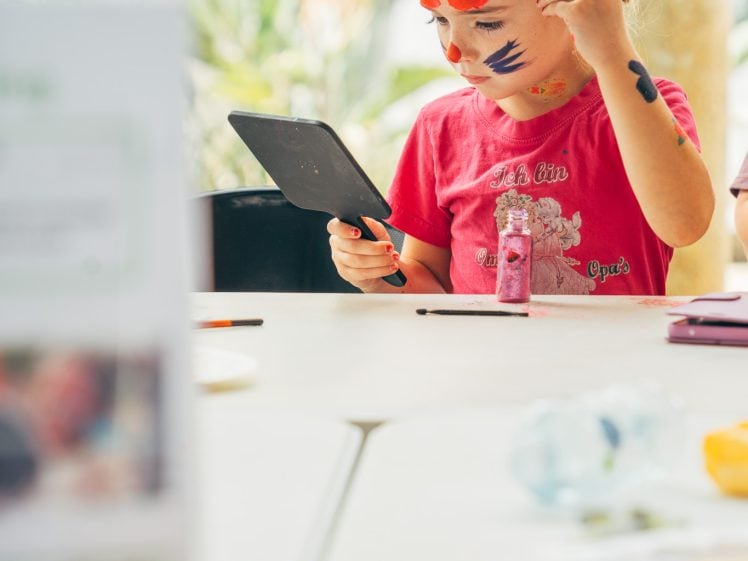 Image of A child wearing bunny ears and face paint looks into a mirror, holding a make-up brush. She sits at a table with art supplies, concentrating on her reflection. The background is bright and slightly blurred.