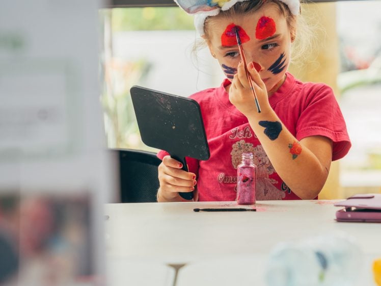 Image of A young child wearing bunny ears paints her face with colourful designs using a brush, while holding a small mirror. She sits at a table with art supplies, and sunlight streams in from a window behind her.
