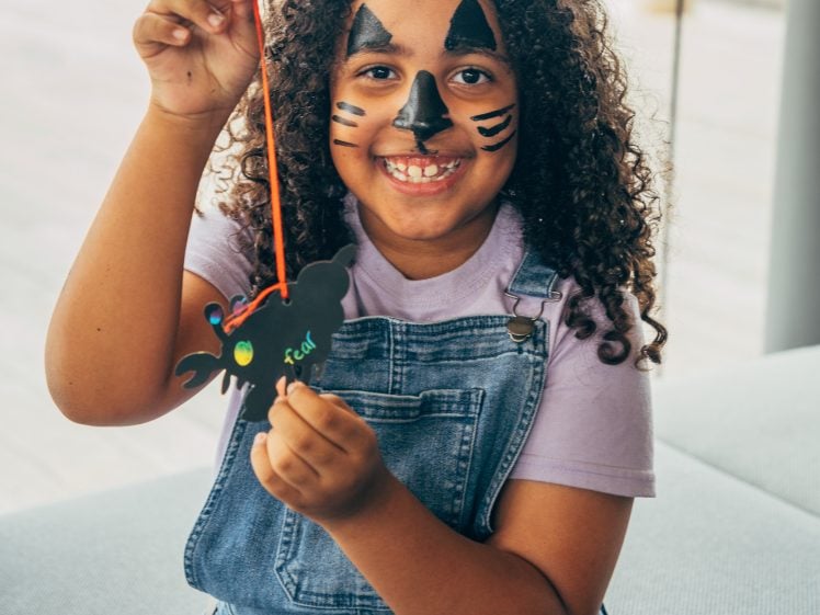 Image of A smiling child with curly hair, wearing dungarees and a t-shirt, has their face painted like a cat and is holding up a black bat-shaped craft on an orange string.