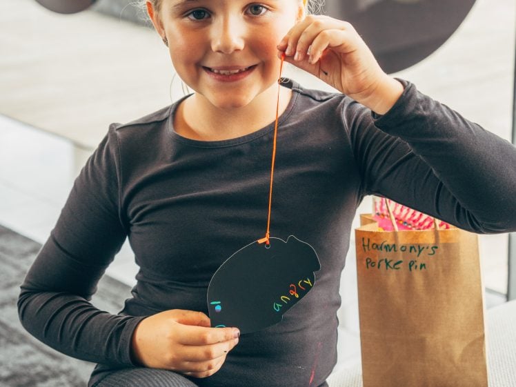 Image of A smiling young girl in a black shirt holds up a decorated black paper cut-out and sits next to a brown paper bag labelled Harmony’s Perky Pin.