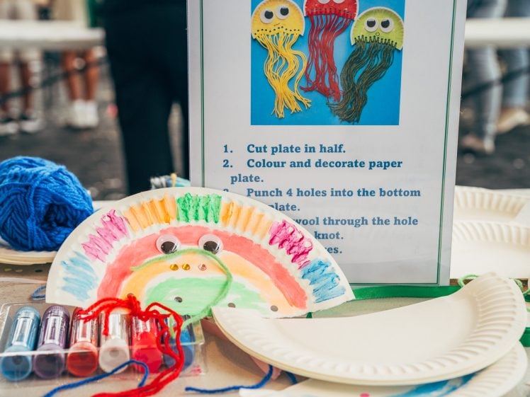 Image of A craft table displays painted paper plate jellyfish with wool tentacles, coloured markers, paint, and a sign with instructions and an example image of three jellyfish. People are gathered in the background.