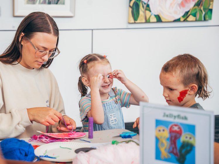 Image of A woman helps two young children with a colourful jellyfish craft at a table. One girl smiles as she holds red craft pieces to her eyes, while a boy with face paint looks on. Art supplies and finished jellyfish art are visible.
