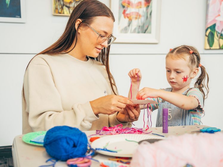 Image of An adult and a young girl with face paint sit at a table doing crafts with colourful yarn, surrounded by art supplies. Paintings hang on the wall behind them.