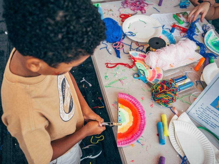 Image of A child stands at a table covered with art supplies, using a felt-tip pen to decorate a colourful paper plate. The table is scattered with wool, felt-tip pens, glue sticks, and other craft materials.